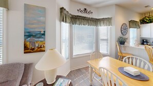 A dining area with a wooden table, white chairs, and a lamp. There is a painting of a beach scene on the wall. - Live the Golf Dream | River Creek Golf Condo in NC | RC 2002 (Sunset Beach)