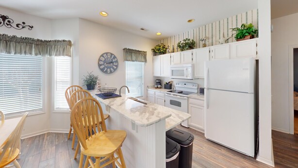 A modern kitchen with white cabinetry, a marble countertop, wooden chairs, a sink, a stove, a microwave, and a refrigerator. - Live the Golf Dream | River Creek Golf Condo in NC | RC 2002 (Sunset Beach)