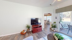 A living room with a flat-screen TV on a wooden stand, a wicker basket with books, a potted plant, and a sliding glass door leading to an outdoor area with a patio table and chairs. - Live the Golf Dream | River Creek Golf Condo in NC | RC 2002 (Sunset Beach)