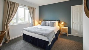 Ein Schlafzimmer mit Bett, Nachttischlampen, einem Sessel und Blick ins Freie durch ein Fenster mit Vorhängen.