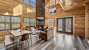 A spacious living area with a fireplace, wooden walls, and large windows. - Luxurious 3BD Lodge with POOL! Hot Tub + Games! (Sevierville)