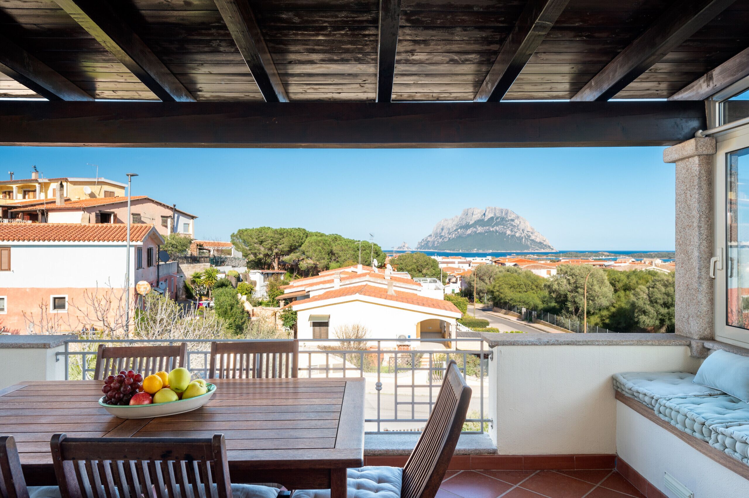 A balcony with a wooden table and chairs, a bowl of fruit, and a view of a coastal town and mountain.