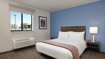 A hotel room with a large bed, a nightstand with a lamp, a window with a view of buildings, and a wall-mounted air conditioner.
