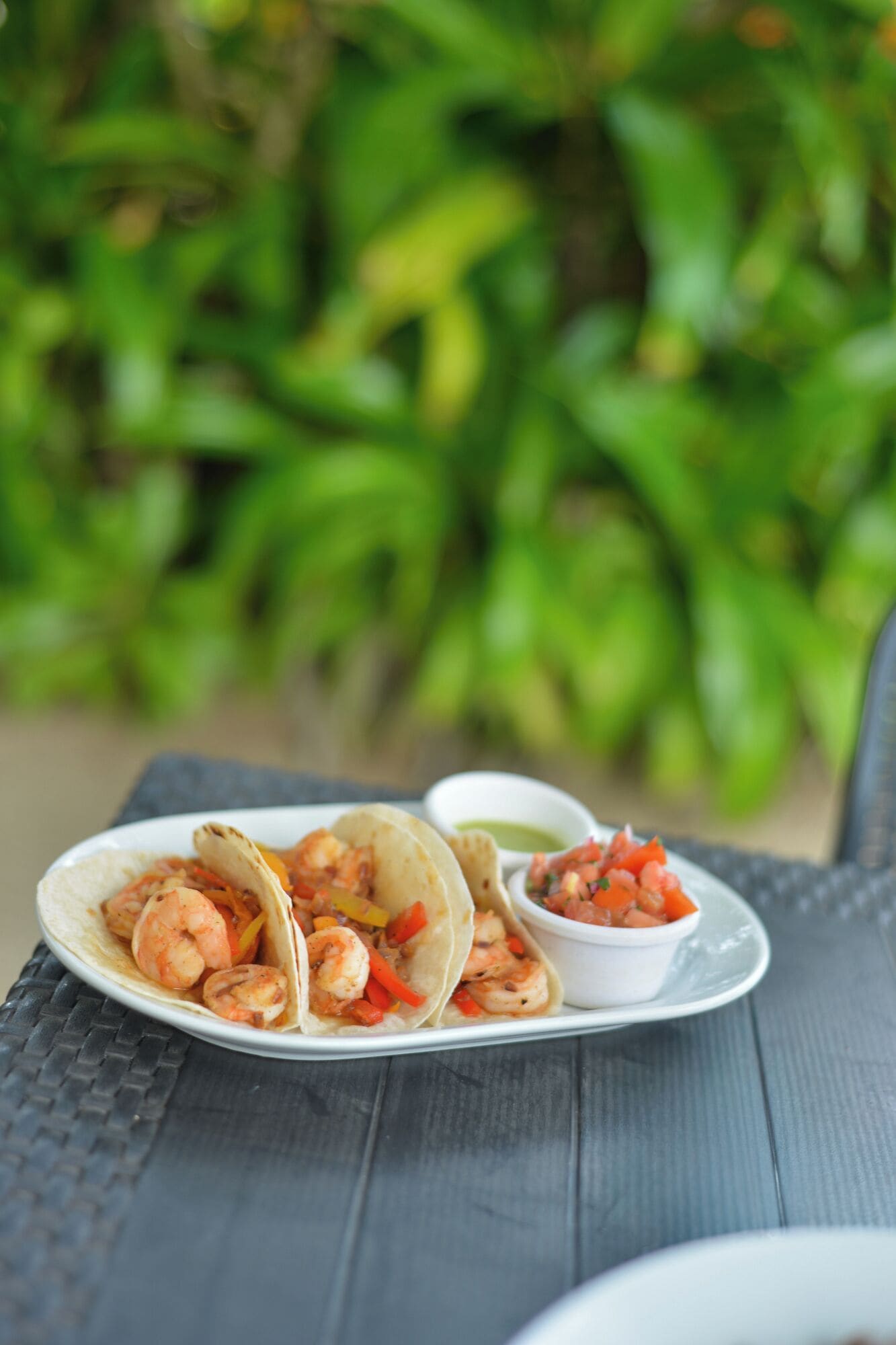 A plate of shrimp tacos with a side of salsa on a table with a green leafy background.