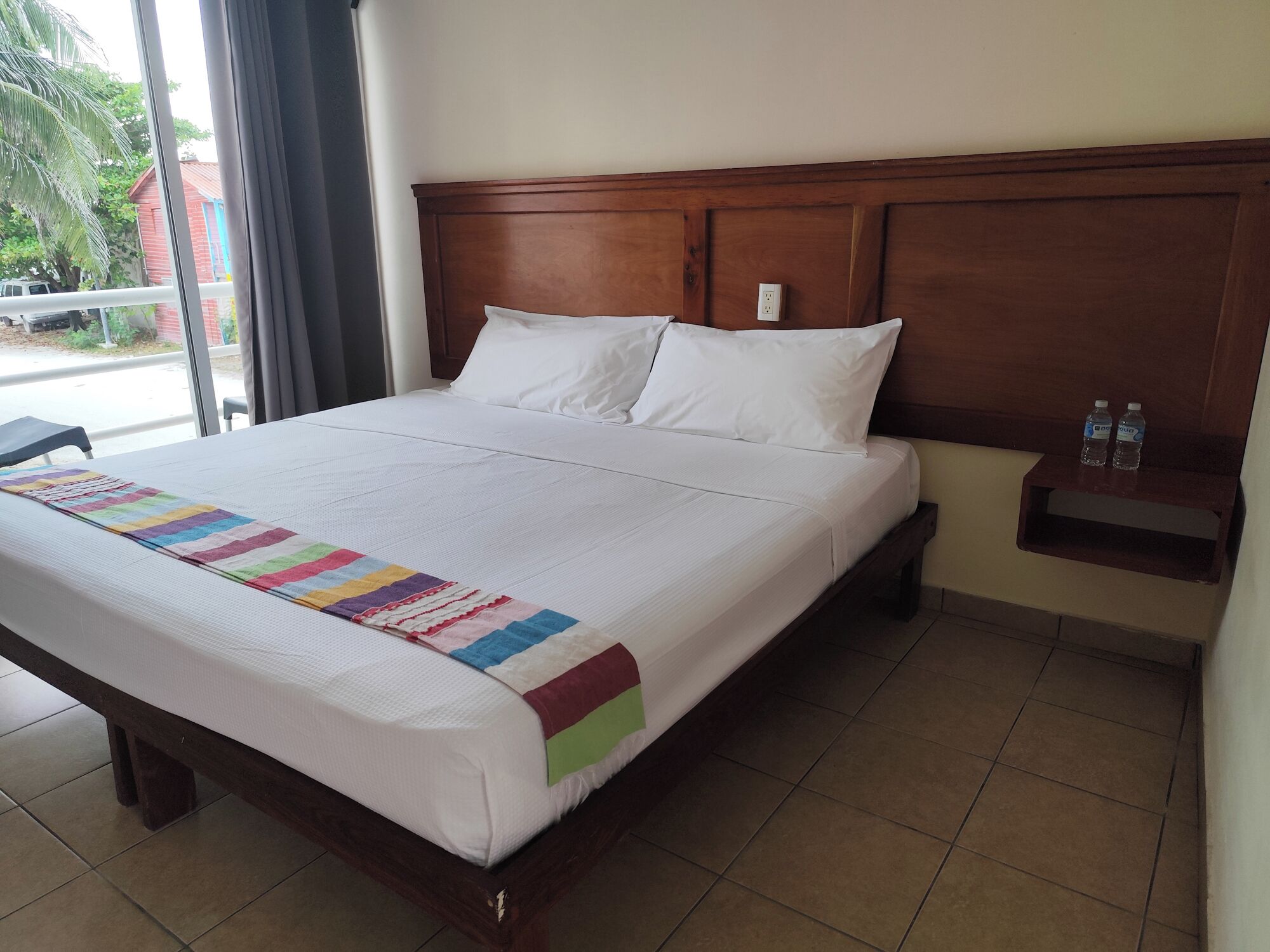 A neatly made bed with a colorful blanket, a wooden headboard, and a small nightstand with water bottles.