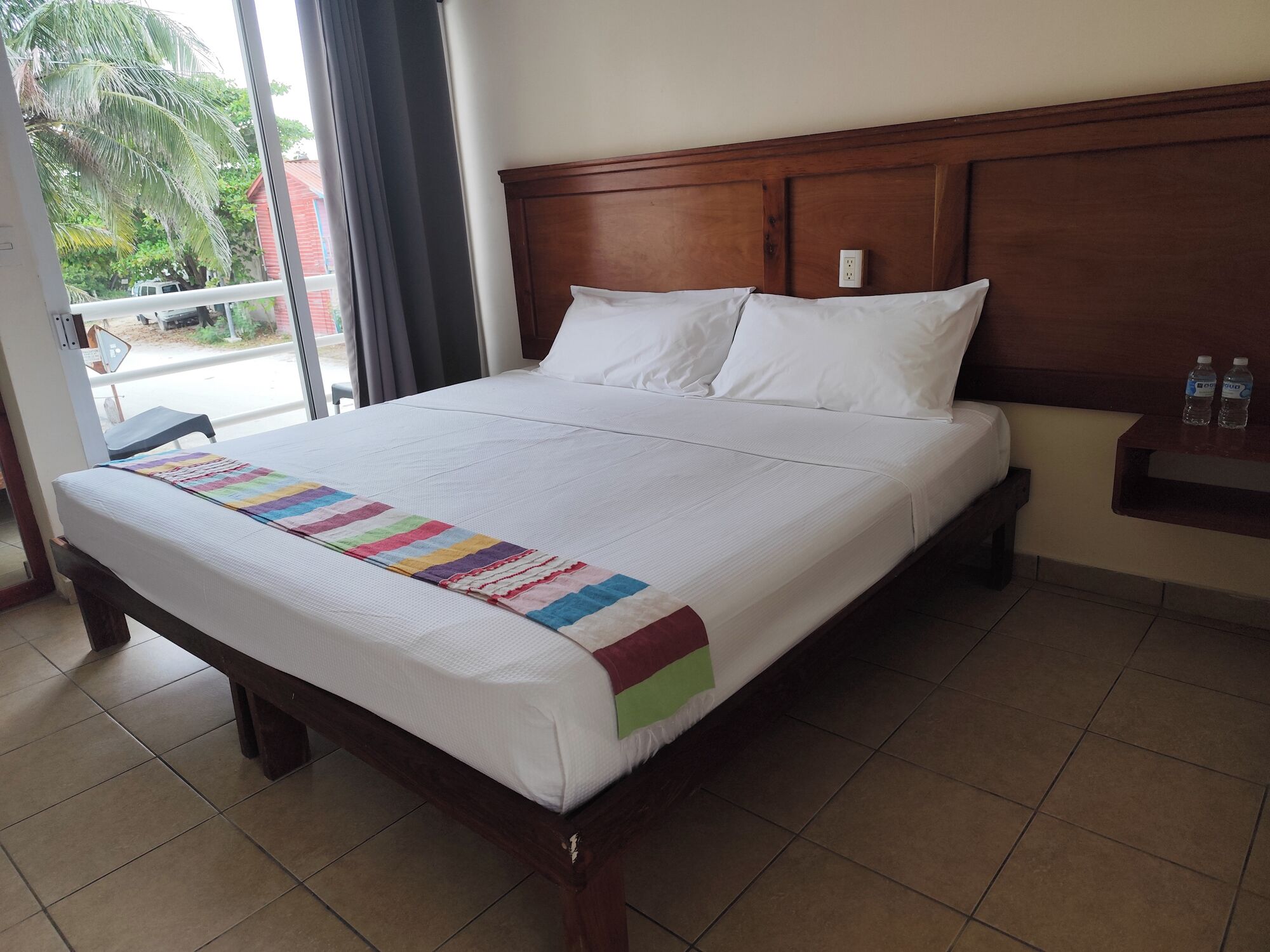 A hotel room with a large bed, a wooden headboard, a window with a view of palm trees, and a small table with water bottles.