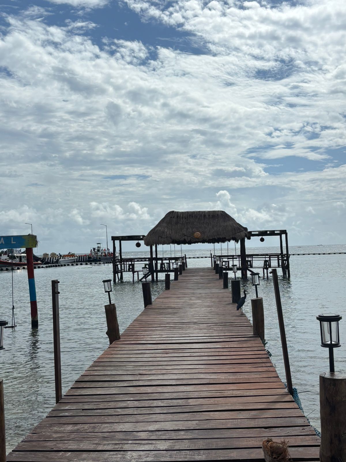 Un muelle de madera que conduce a una estructura con techo de paja sobre el agua, con cielo despejado y barcos a lo lejos.