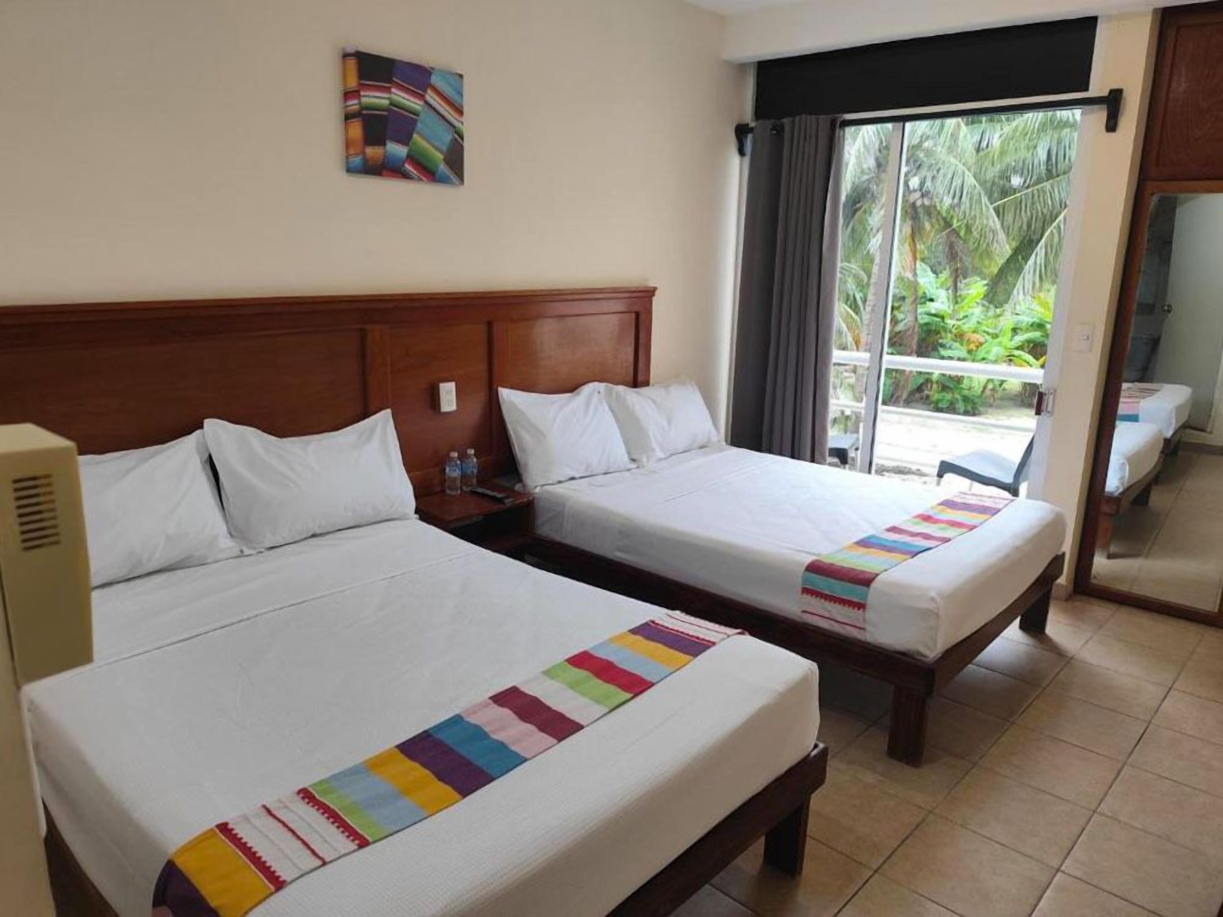 A hotel room with two beds, a wooden headboard, a window with curtains, and a view of greenery outside.