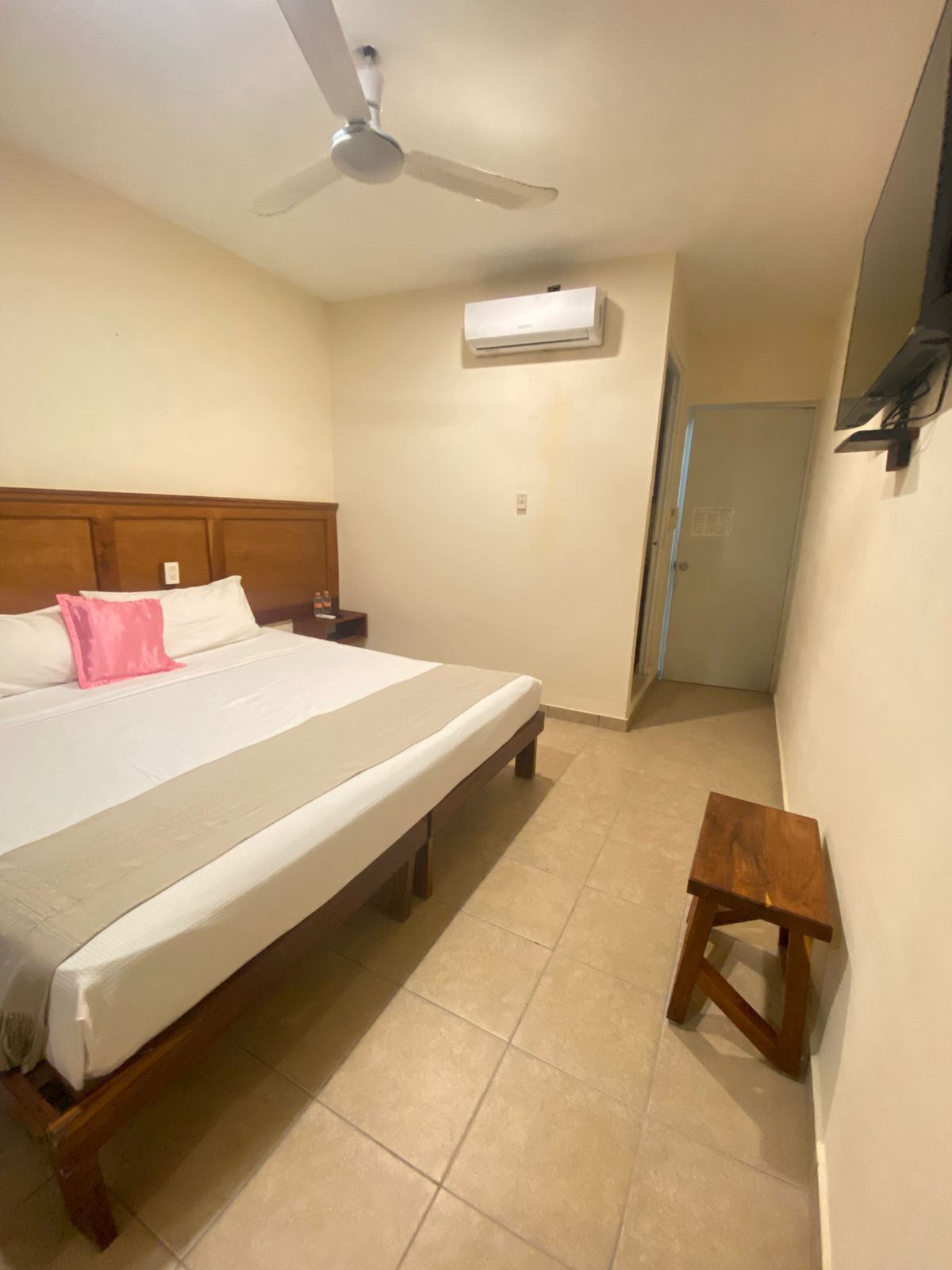Un dormitorio con una cama, ventilador de techo, aire acondicionado, mesita de noche y un taburete de madera.