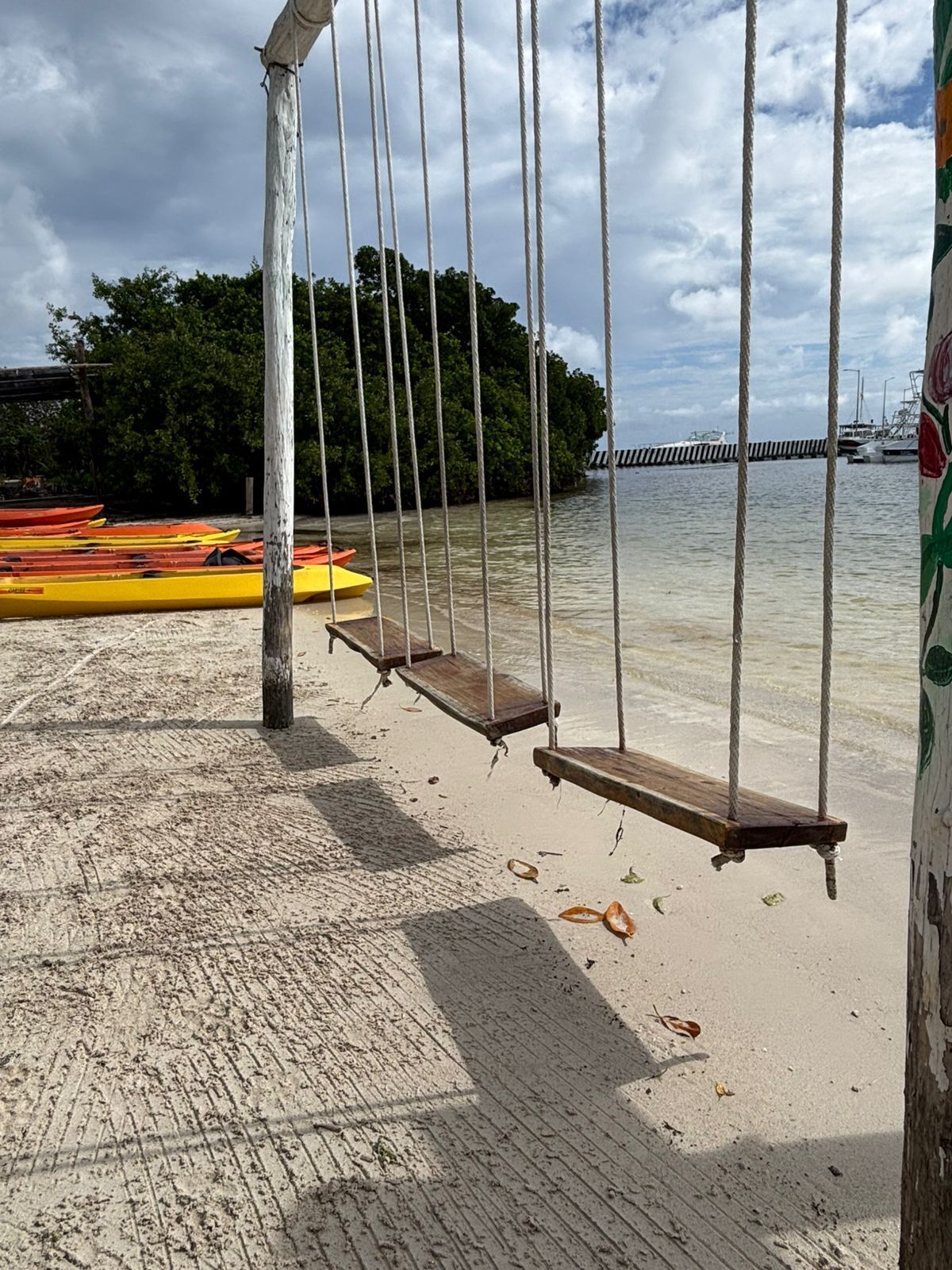A sandy beach with a swing set, kayaks, and a dock with boats in the background.