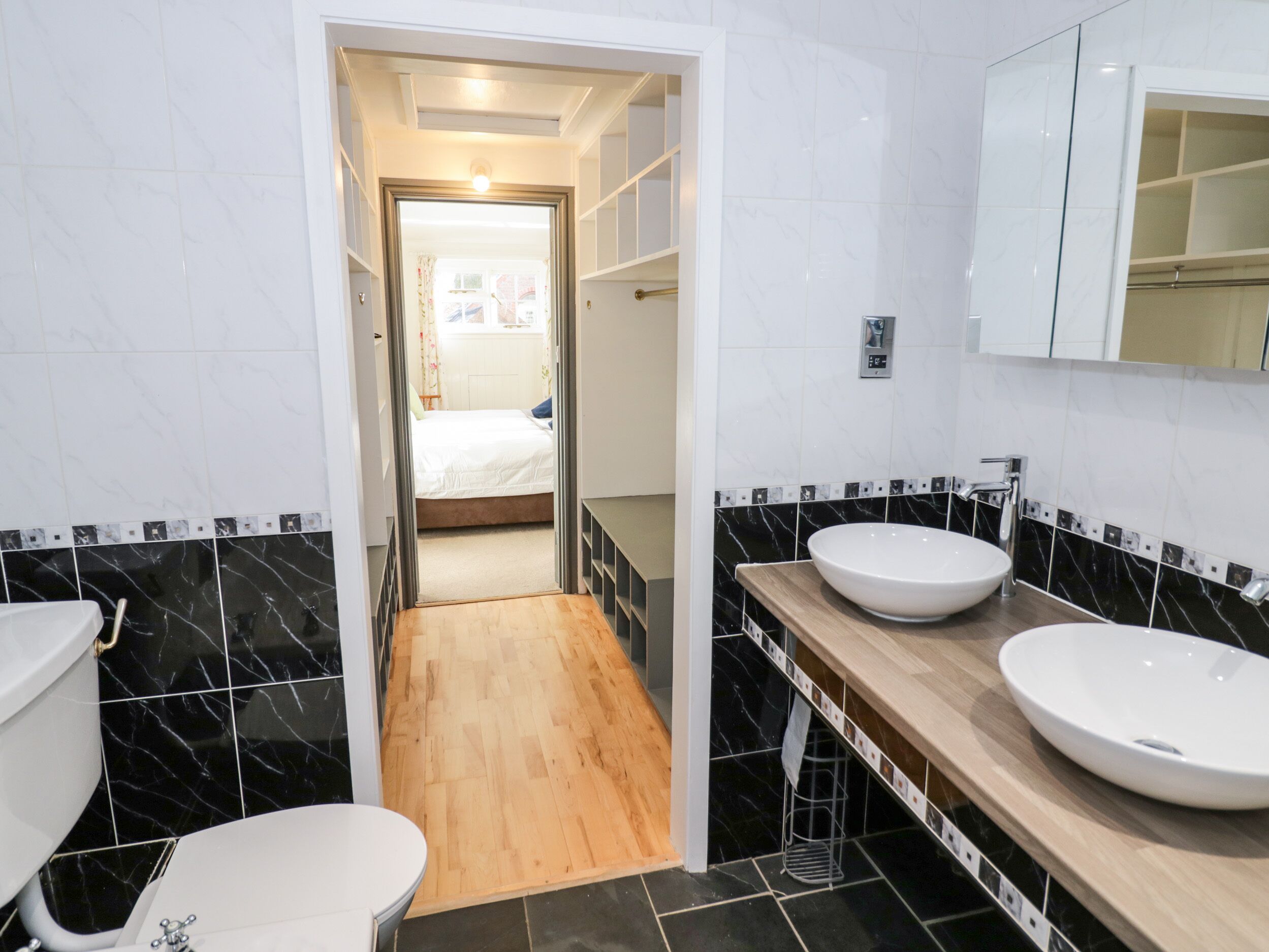 A modern bathroom with two sinks, a toilet, and a wooden floor leading to a bedroom.
