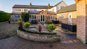 Une maison à deux étages avec une façade en briques, une porte d’entrée et un petit jardin agrémenté de plantes en pot et d’une table de patio.