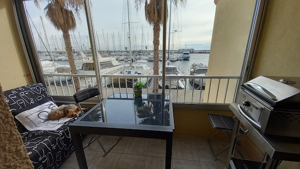 A balcony with a view of a marina, a small table with a plant, a grill, and a sofa with a dog. - Studio Trés Cocooning Pour Couple Aimant la Tranquillité, Avec Enfants (Gruissan)