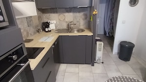 A compact kitchen with grey cabinets, a stainless steel sink, a built-in oven, and a small dining table with a patterned tablecloth. - Studio Trés Cocooning Pour Couple Aimant la Tranquillité, Avec Enfants (Gruissan)