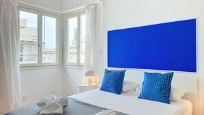 Ein Hotelzimmer mit Bett, weißen Vorhängen, einer blauen Akzentwand und Blick auf Gebäude.