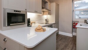 A modern kitchen with white cabinetry, a built-in oven, a coffee maker, and a wooden cutting board with a knife. - Quarters (Ambleside)