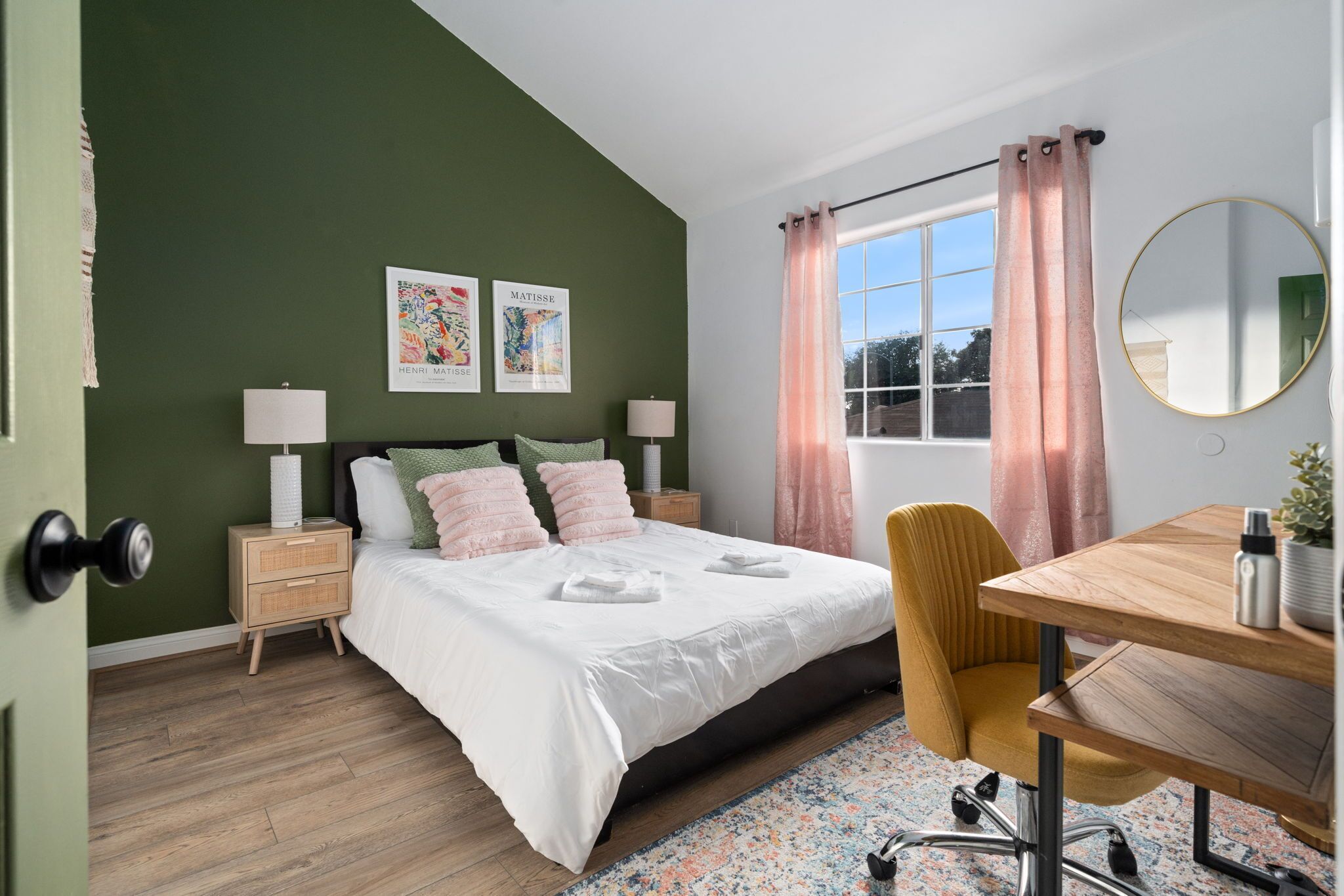 A bedroom with a bed, bedside tables, a desk, a chair, a window with curtains, and wall art.