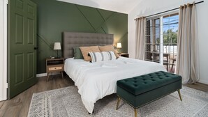A bedroom with a large bed, a green upholstered bench, a nightstand with a lamp, and a view through a window with curtains.