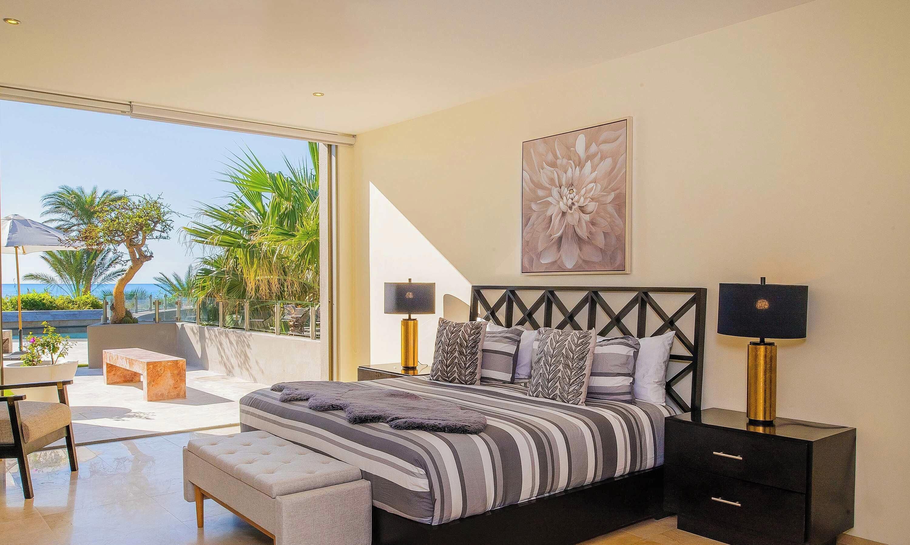 A modern bedroom with a large bed, two bedside tables, and a view of palm trees and the sea.