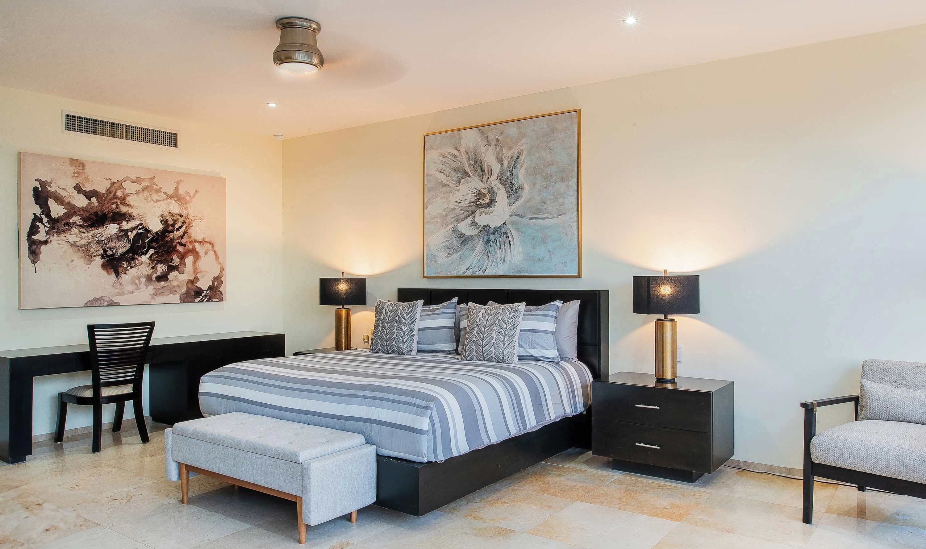A modern bedroom with a large bed, two bedside tables with lamps, a chair, and two large abstract paintings on the walls.