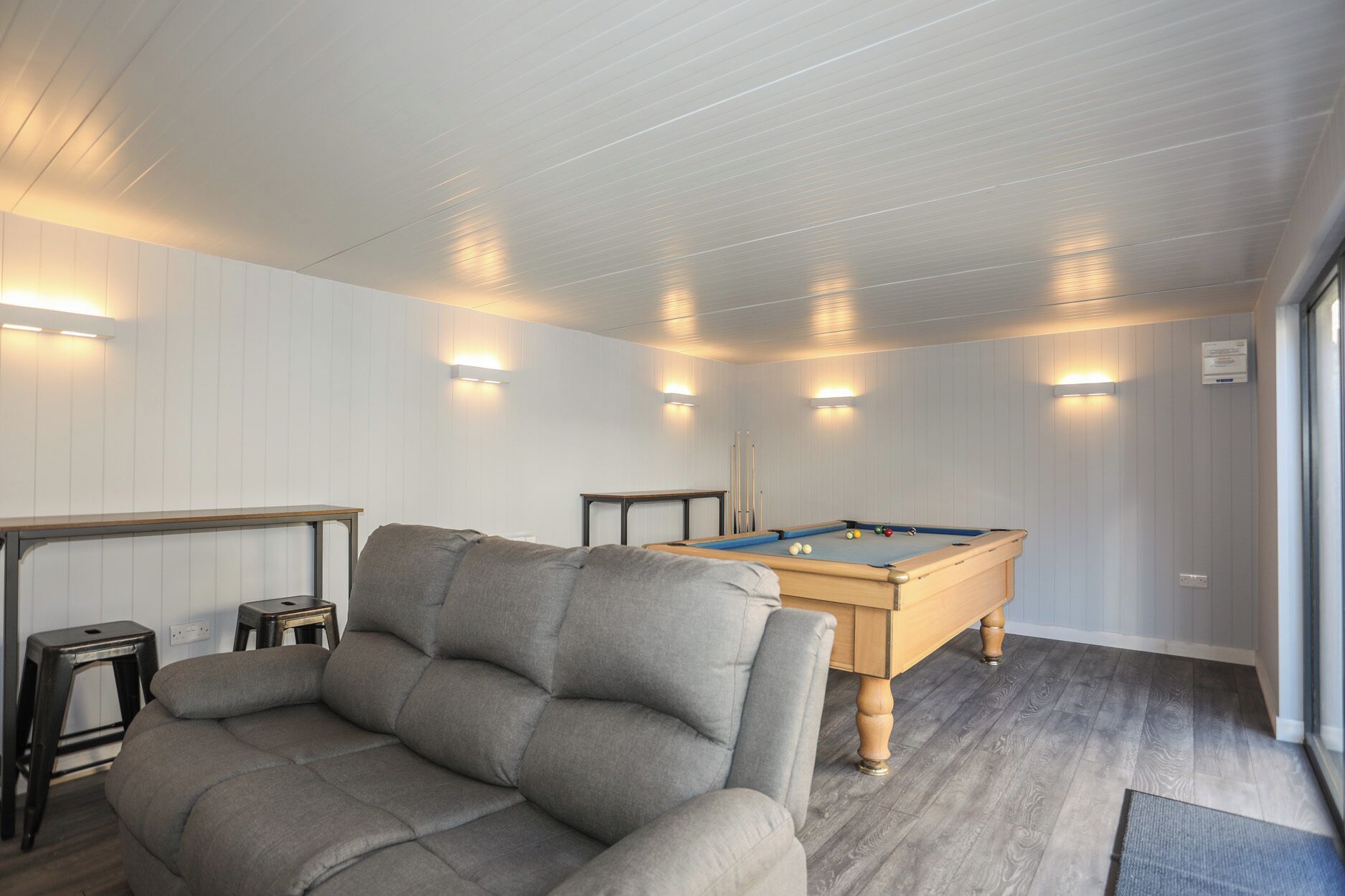 A pool table in a room with white paneled walls and ceiling, two small tables, and a grey sofa.