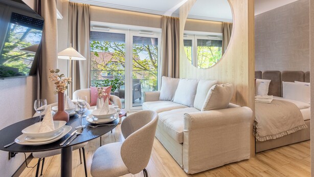 Een moderne hotelkamer met een bank, een eettafel met borden en glazen, een vaas met bloemen en een grote spiegel.