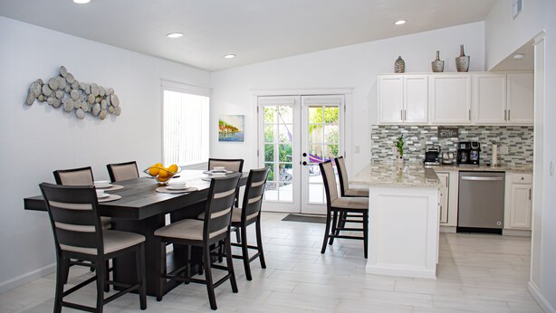 A modern kitchen with white cabinetry, a central island, and a dining area with a dark wooden table and chairs. - Coachella Poolside Paradise, accommodates up to 10 guest (Indio)