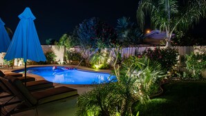 A swimming pool with lounge chairs and umbrellas, surrounded by tropical plants and a wooden fence. - Coachella Poolside Paradise, accommodates up to 10 guest (Indio)