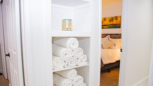 A hotel room with a neatly organized shelf holding rolled towels, a painting on the wall, and a bed with white bedding. - Coachella Poolside Paradise, accommodates up to 10 guest (Indio)