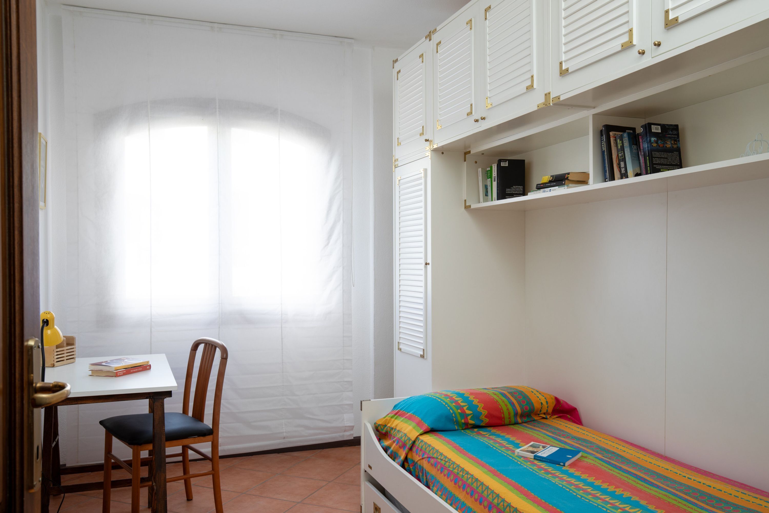 A bedroom with a bed, a desk, and a chair. There is a window with blinds, a bookshelf, and a door.