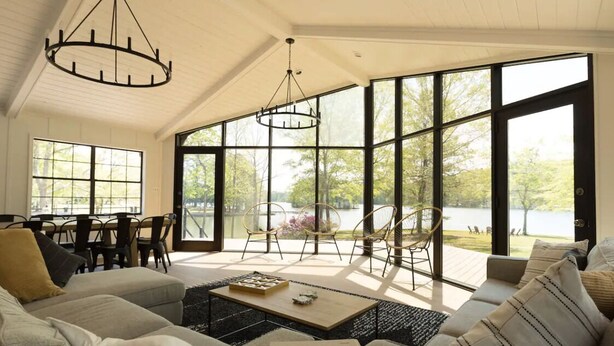 Un vaste salon doté d’une grande fenêtre donnant sur un lac, un coin repas avec une table et des chaises, et un espace salon confortable avec un canapé et des coussins.