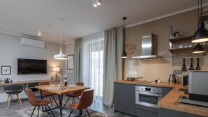 Et moderne køkken med en central ø, indbygget ovn og stålstålelementer. Der er et spiseområde med et træbord og stole, samt et stort vindue med lette gardiner.
