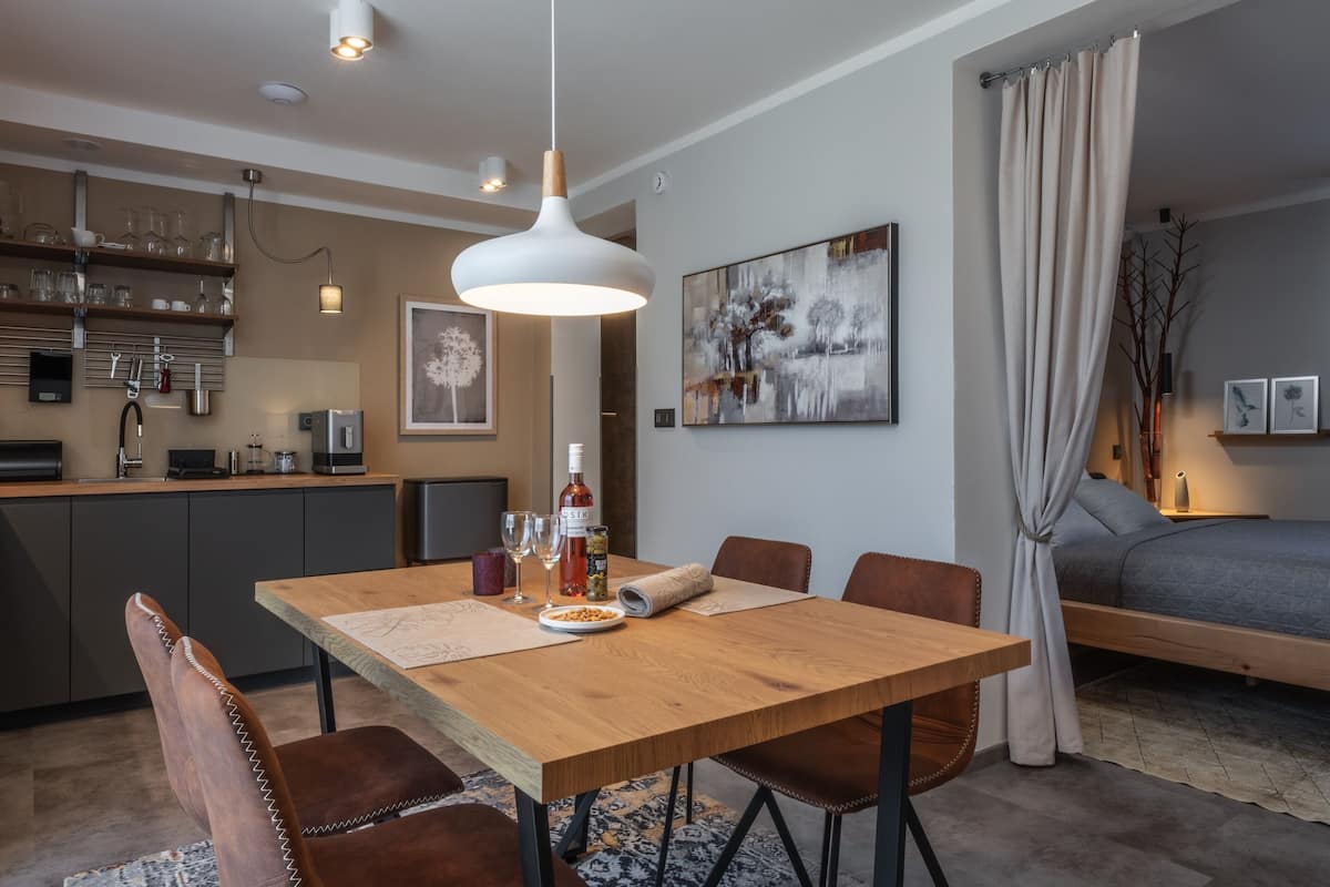Ein moderner Essbereich mit einem Holztisch, Stühlen und einer Pendelleuchte. Im Hintergrund ist eine Küche mit Geräten und einer Bar zu sehen.