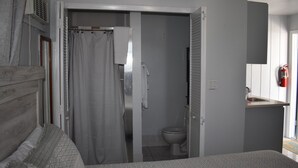 Una camera da letto con un letto, un bagno con il water e tenda da doccia, e un angolo cottura con lavello e estintore.