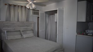 Una camera da letto con testiera in legno, un letto con biancheria a motivi, un ventilatore a soffitto, un bagno con tenda doccia e un angolo cottura con lavello e una bottiglia.