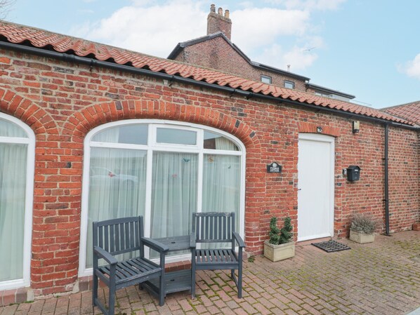 A brick cottage with a white door, two windows with white curtains, and a small garden with a bench and potted plants.
