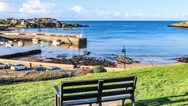 A coastal view with a bench overlooking a harbor, boats, and a distant town. - Ty Bara (Cemaes Bay)