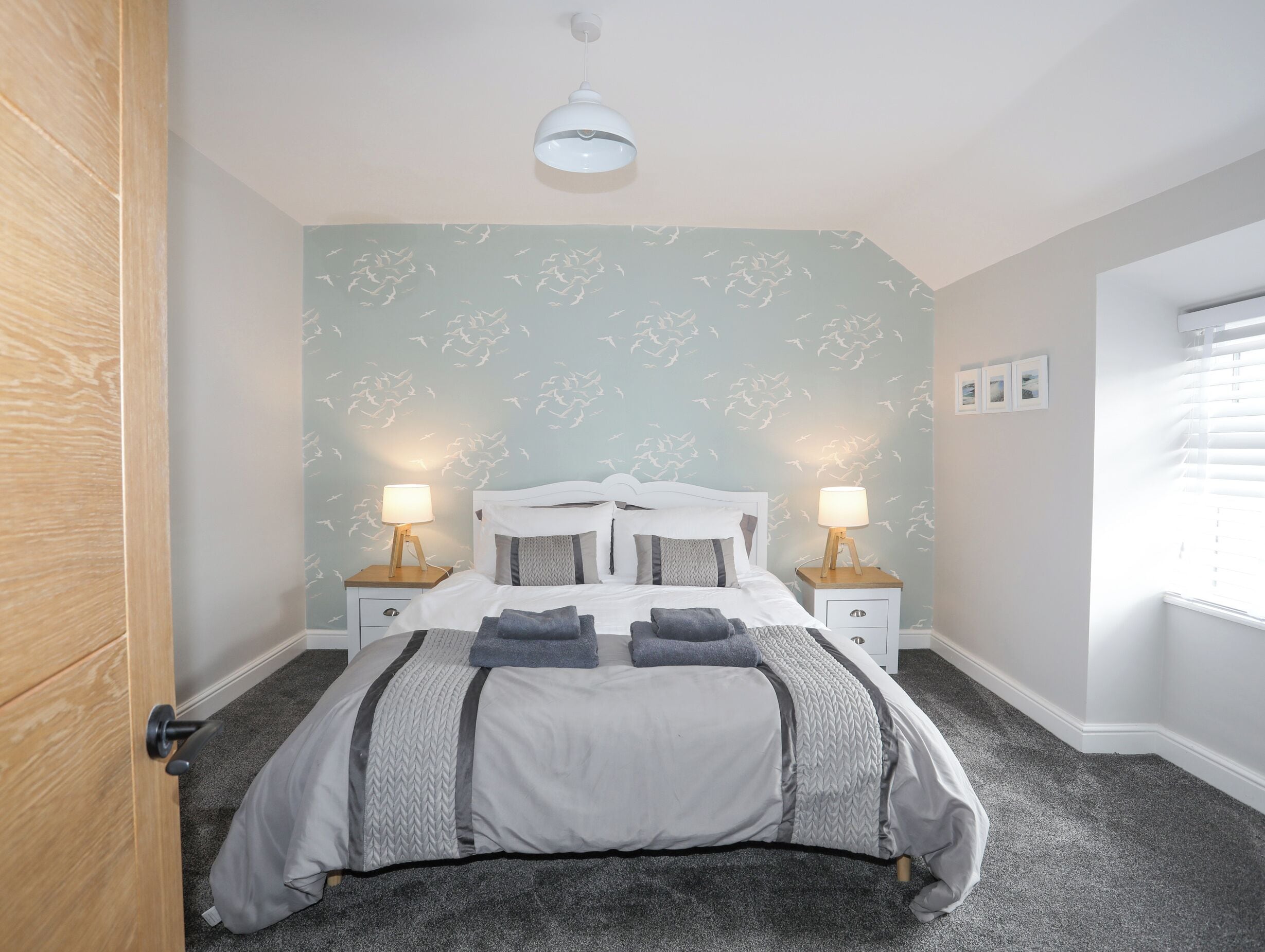 Une chambre à coucher avec un lit, deux tables de chevet avec des lampes, une porte en bois et une fenêtre avec des stores.