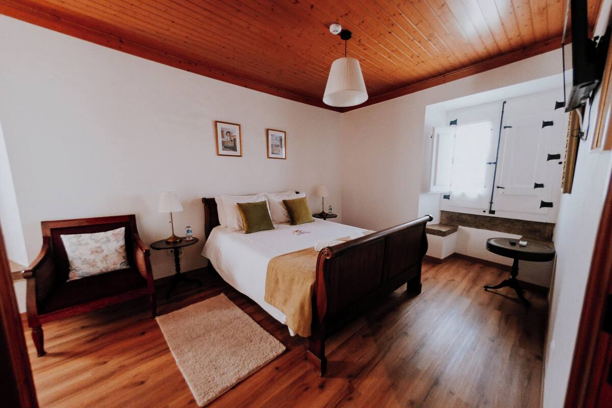 A bedroom with a wooden ceiling, a bed with pillows, a bedside table, a chair, a window, and a small table.