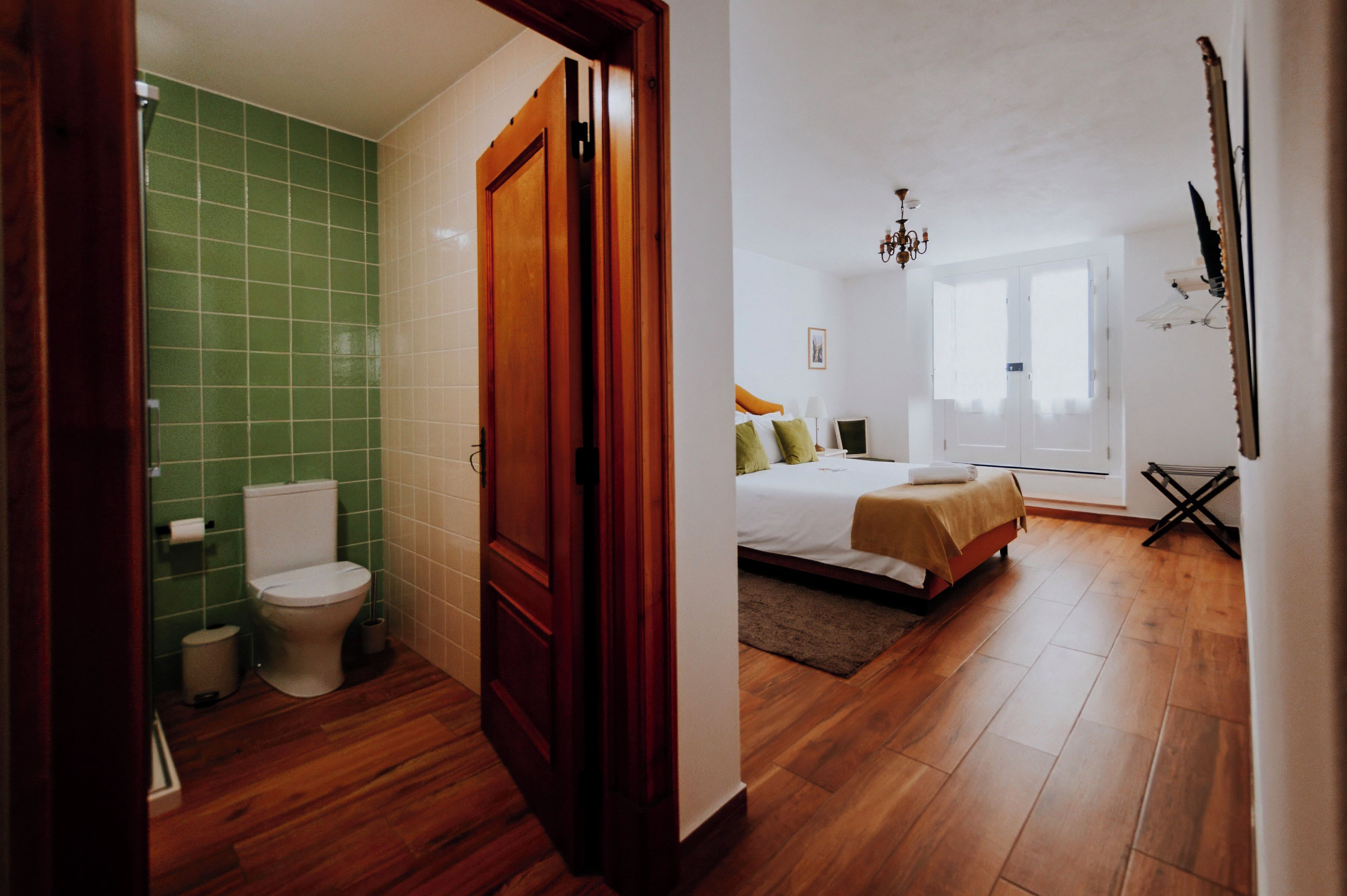 A bathroom with a toilet, sink, and green tiles, and a bedroom with a bed, window, and wooden floor.