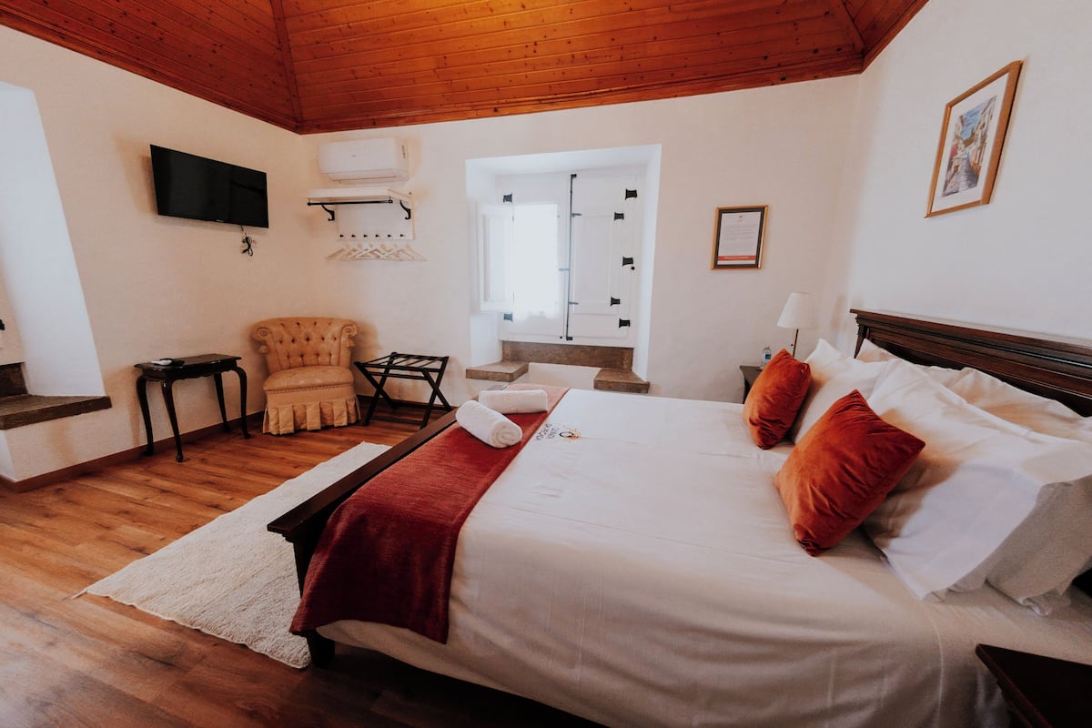 A bedroom with a wooden ceiling, a bed with white linens and red pillows, a chair, a small table, a TV mounted on the wall, and a window with blinds.