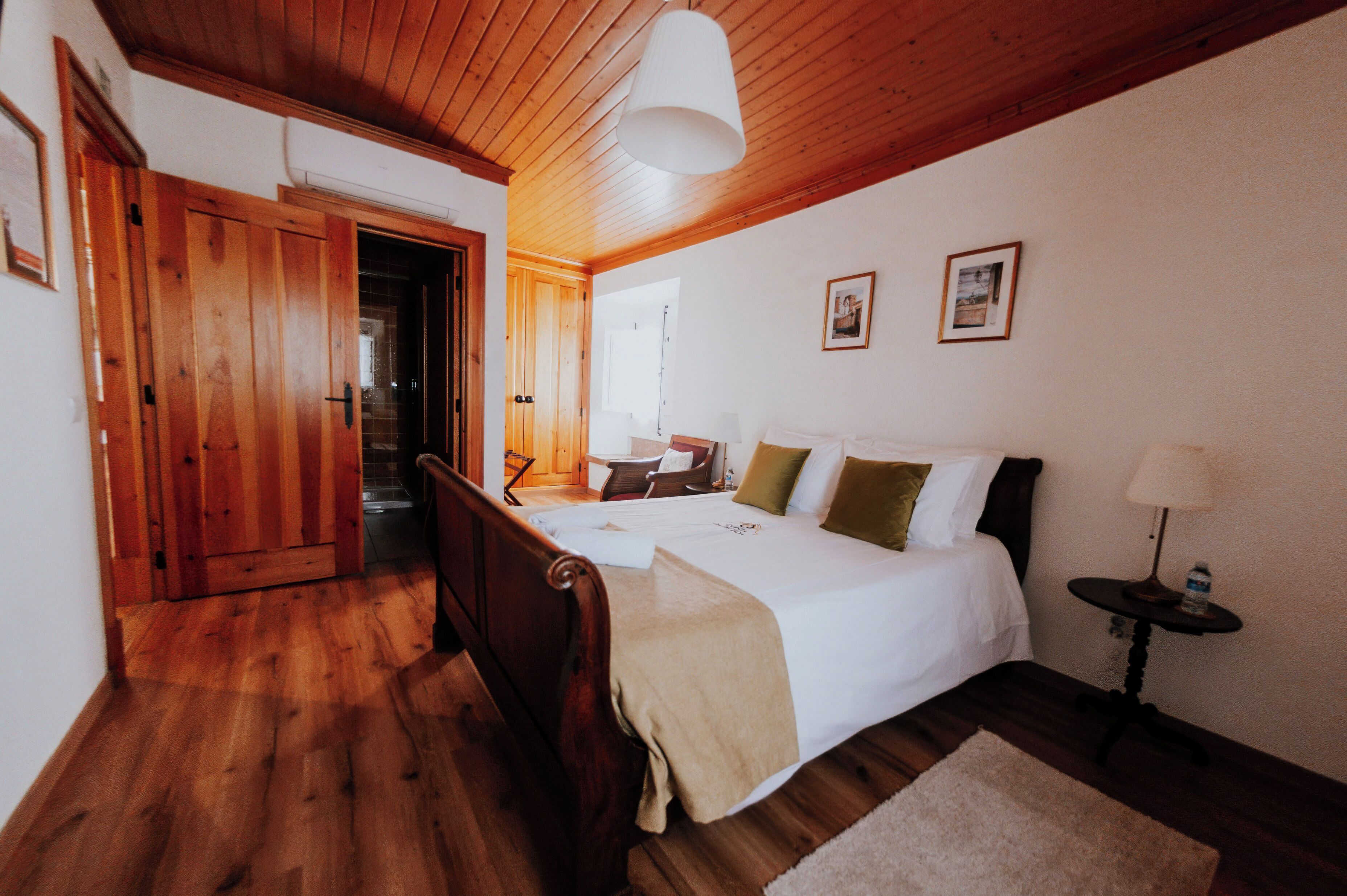 A bedroom with a wooden floor, a bed with white bedding, a bedside table with a lamp, and a door leading to another room.