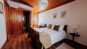 A bedroom with a wooden floor, a bed with white bedding, a bedside table with a lamp, and a door leading to another room.