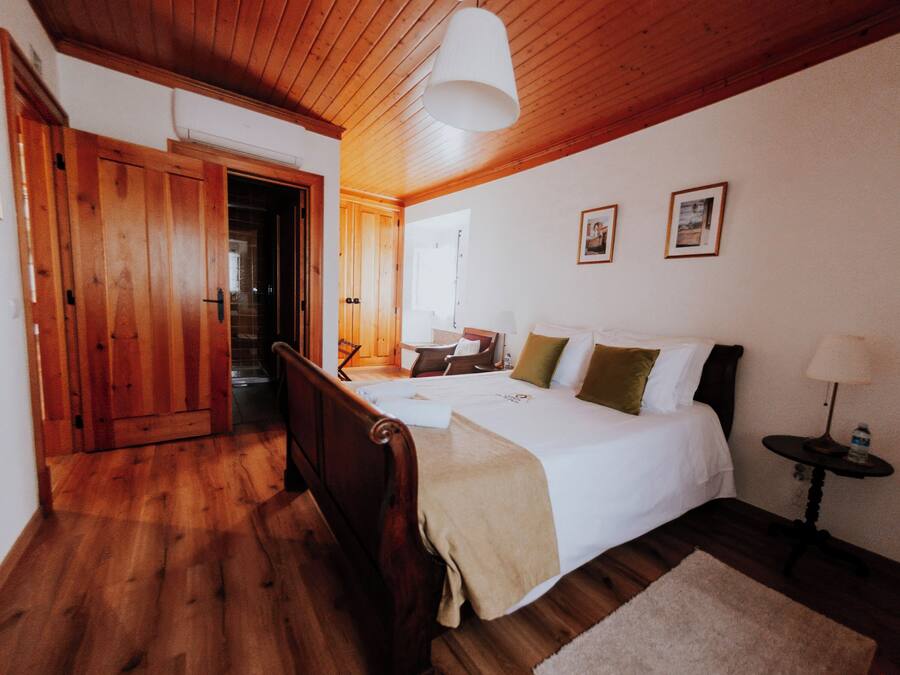 A bedroom with a wooden floor, a bed with white bedding, a bedside table with a lamp, and a door leading to another room.