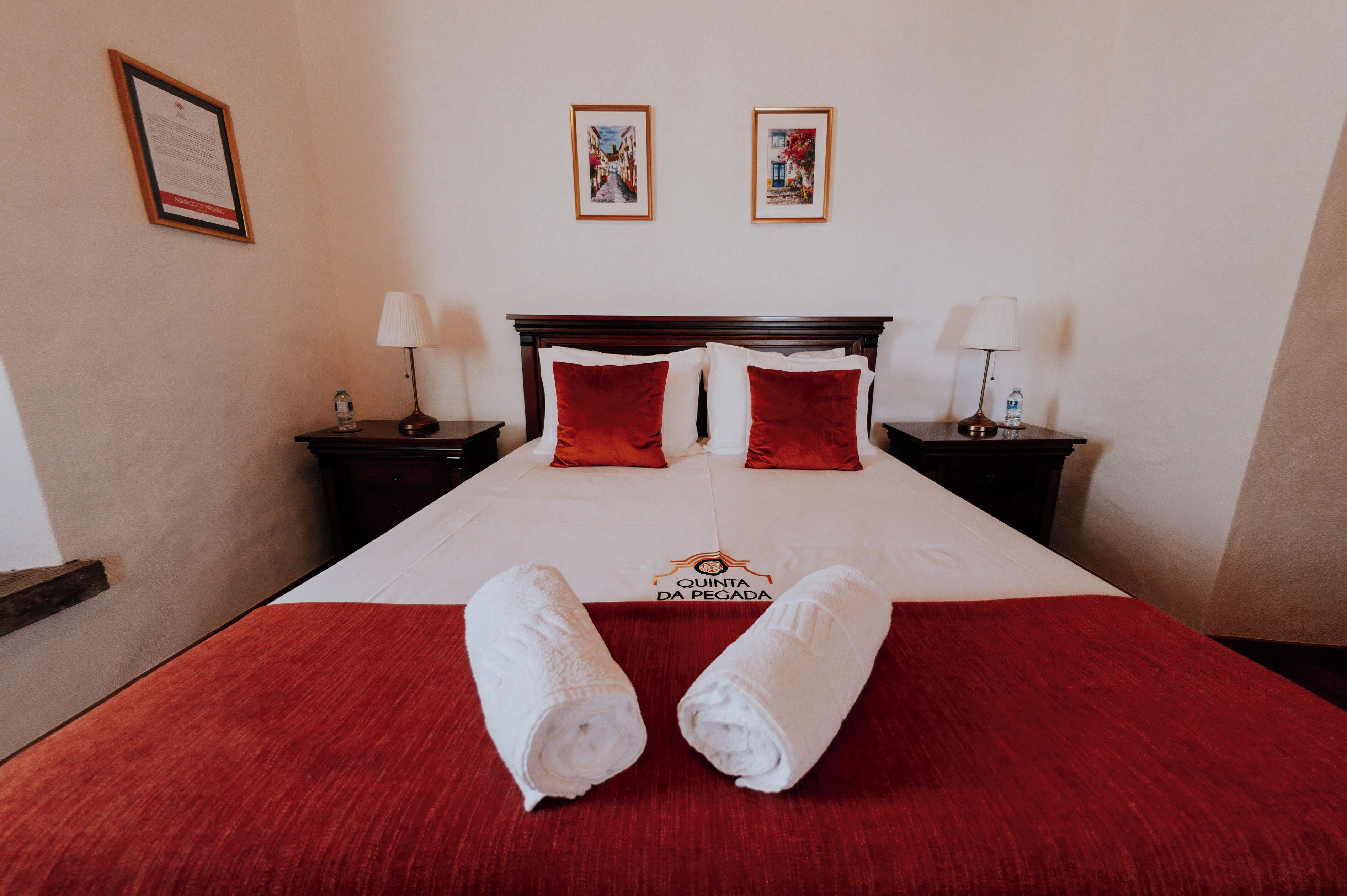 Habitación de hotel con cama, dos mesitas de noche, colcha roja, dos almohadas rojas, dos toallas blancas y dos cuadros en la pared.