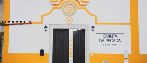 A building facade with a yellow and white color scheme, featuring a large black door and a sign that reads "QUINTA DA PEGADA Casa de Campo."