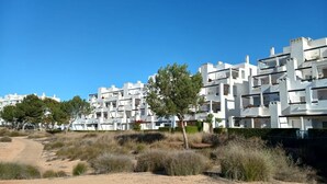 A modern apartment complex with multiple floors and balconies, surrounded by dry vegetation and a clear blue sky.