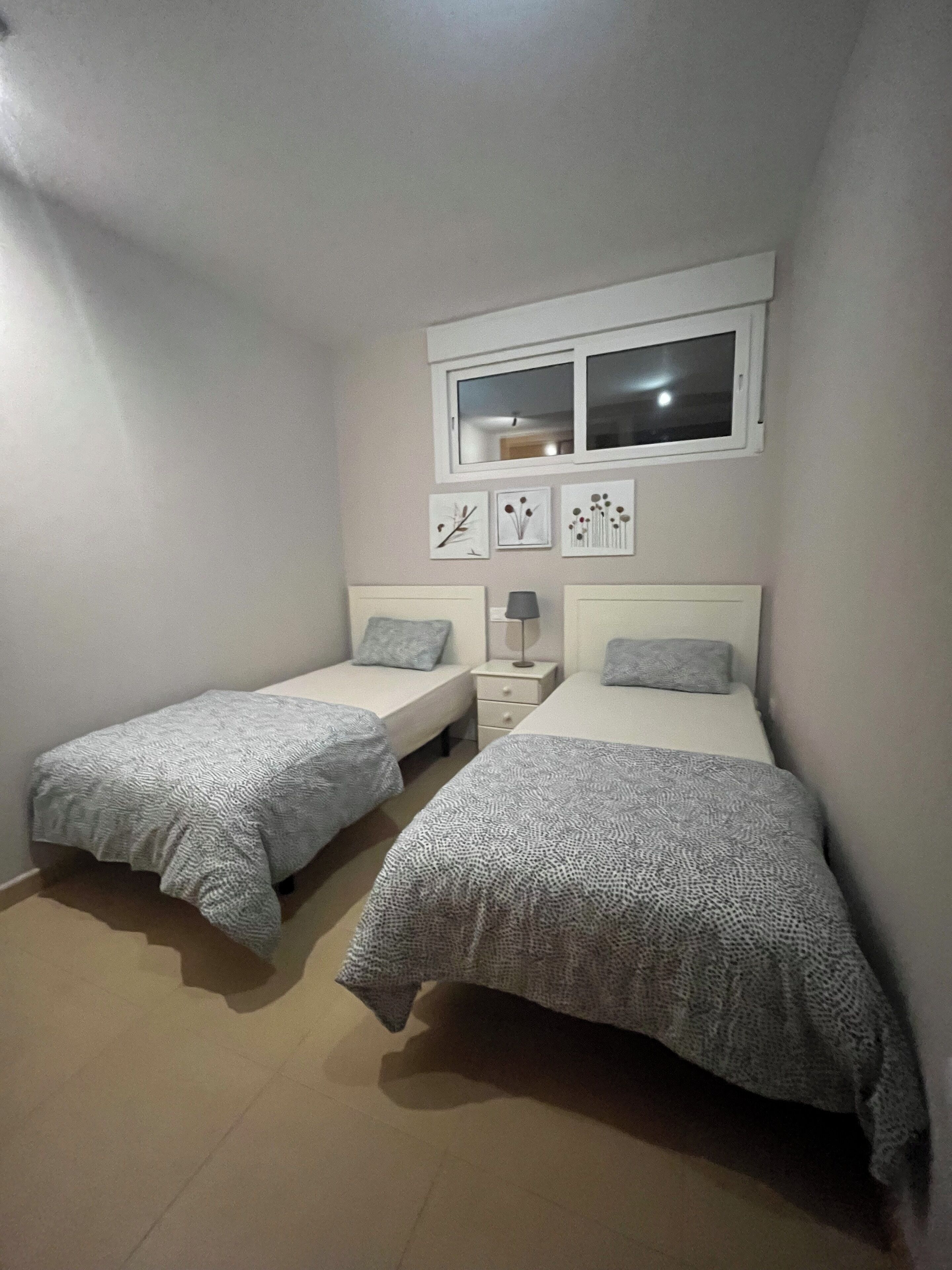 A small, well-lit bedroom with two single beds, a bedside table with a lamp, and a window above the beds.