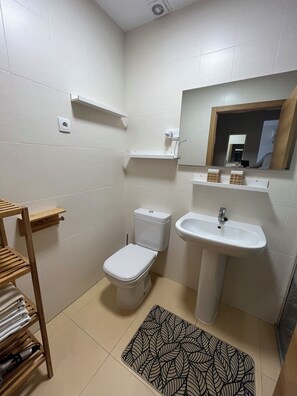 Un baño pequeño y limpio con inodoro, lavabo y espejo. Hay estantes sobre el inodoro y el lavabo, y un perchero de madera con toallas.