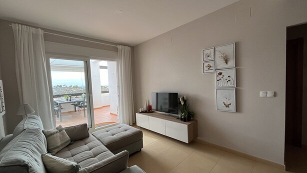 A modern living room with a sectional sofa, a flat-screen TV on a white console, and a balcony with a view.